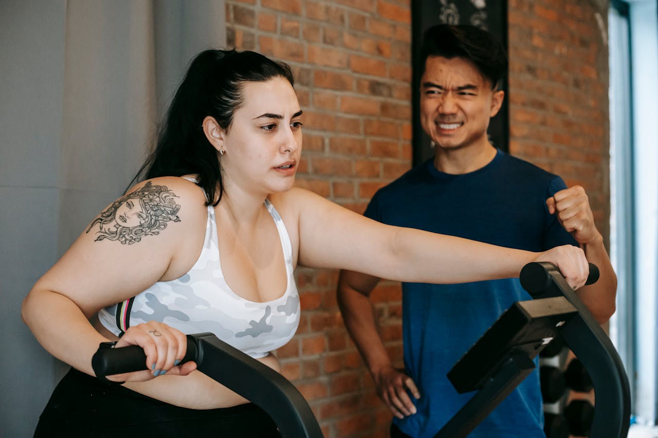 How Women Working in Demanding Careers Can Make Time for Physical Wellness? Image of a woman exercising on machine in gym with trainer encouraging her, brick wall background behind them.