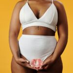 Image of a woman in white wear holding half of a grapefruit in front of her pelvis, on orange background, showing how to keep your vagina clean and smelling good.
