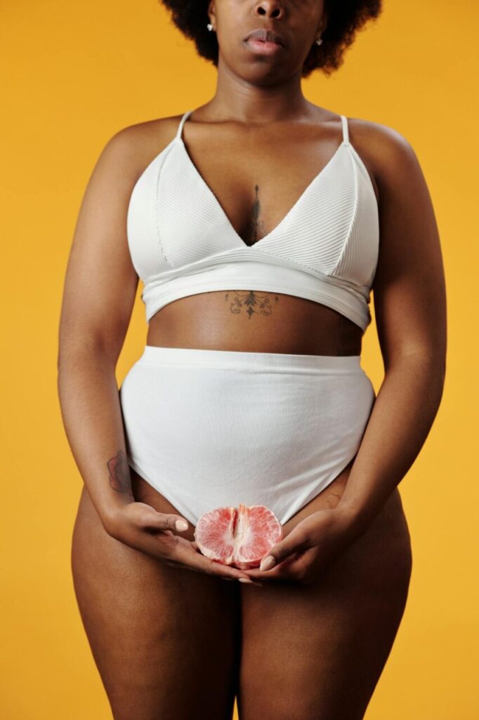 Image of a woman in white wear holding half of a grapefruit in front of her pelvis, on orange background, showing how to keep your vagina clean and smelling good.