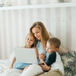 Image of a smiling mother sits on bed with two young children, watching something together on a laptop, showing how to survive being a stay at home mom financially.