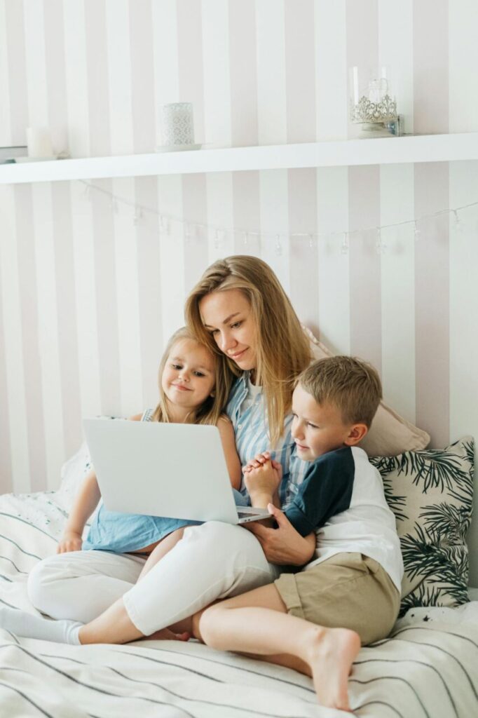 Image of a smiling mother sits on bed with two young children, watching something together on a laptop, showing how to survive being a stay at home mom financially.