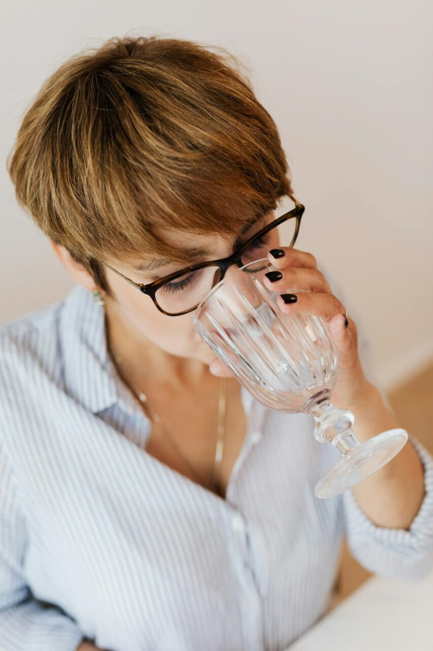 How to Keep Your Vagina Clean and Smelling Good? Image of a woman with short brown hair and glasses drinking water from a clear glass while wearing a striped shirt.