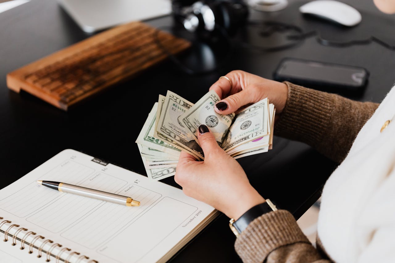 How to Survive Being a Stay at Home Mom Financially? Close-up image of a woman counting U.S. dollar bills at a desk with a notebook, pen, and computer nearby.