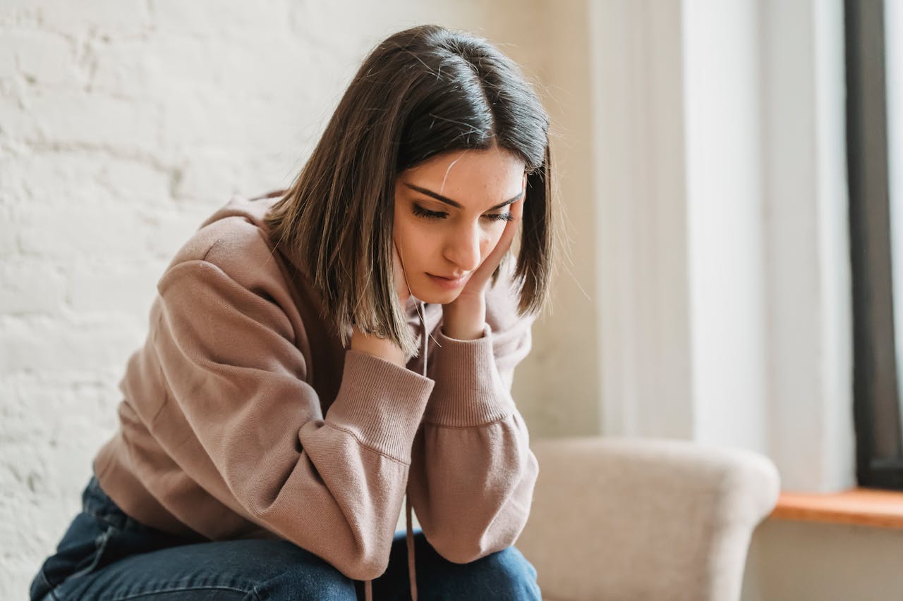 Image of a young woman sitting indoors, looking down sadly with hands pressed to her face, representing can you get depressed without having depression.