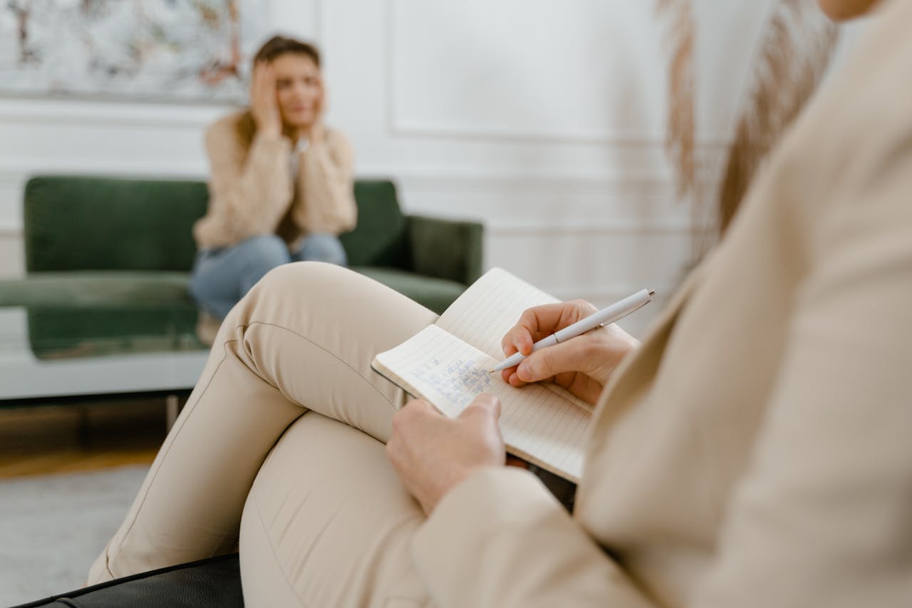 Image of a therapist taking notes during a session with a distressed woman sitting on a green couch in the background.