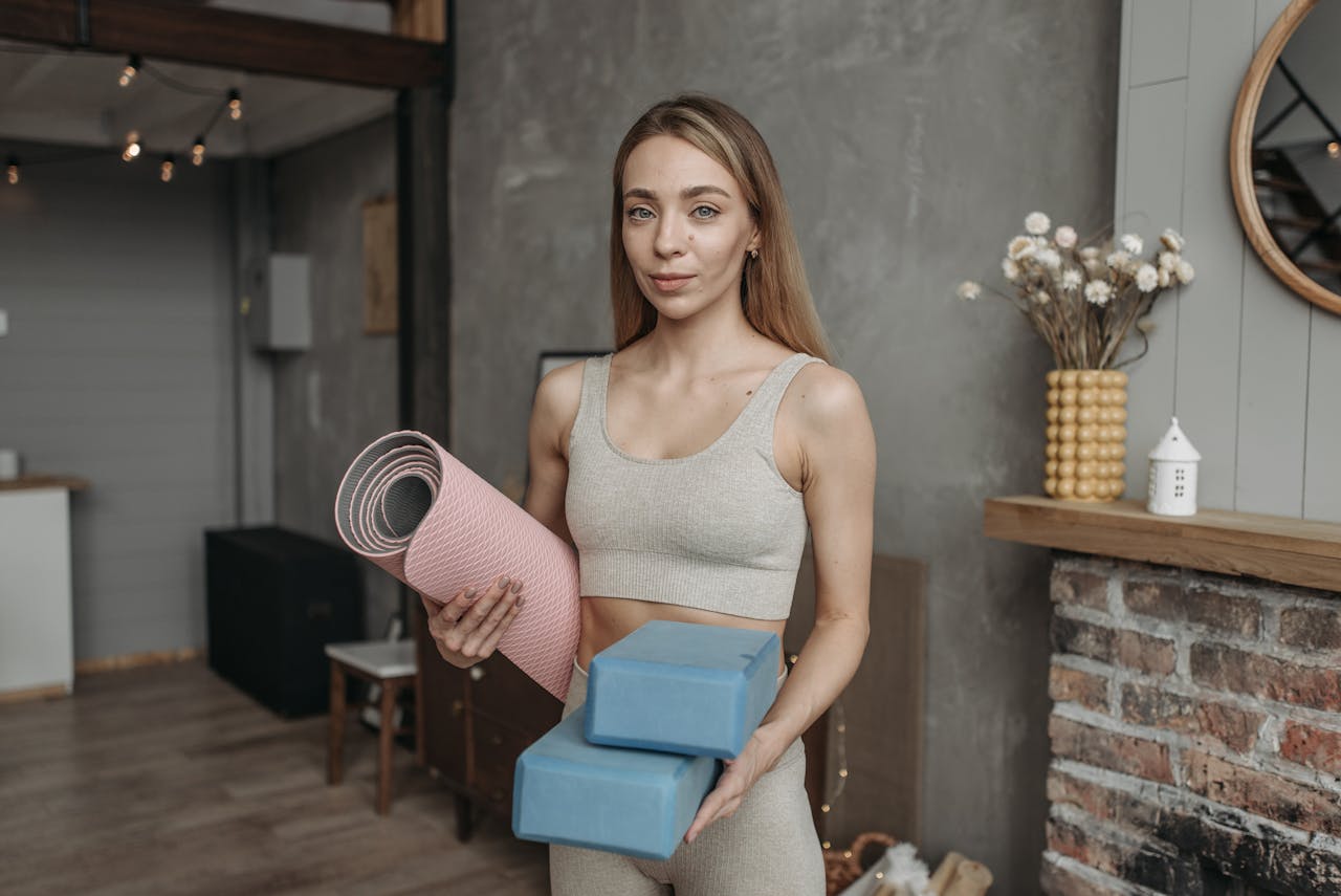 How Women Working in Demanding Careers Can Make Time for Physical Wellness? Image of a fit woman holding a yoga mat and two yoga blocks, standing in a cozy, rustic home environment.