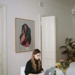 Image of a woman working on a laptop at a white desk in a stylish room with art and flowers in a vase, showing how women working in demanding careers can make Time for physical wellness.