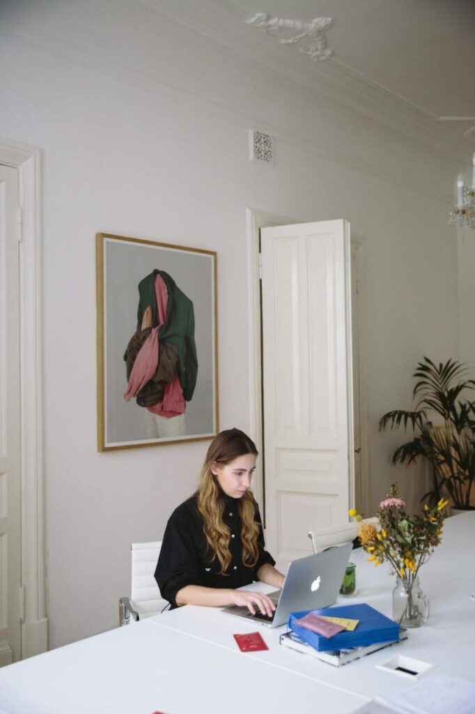 Image of a woman working on a laptop at a white desk in a stylish room with art and flowers in a vase, showing how women working in demanding careers can make Time for physical wellness.