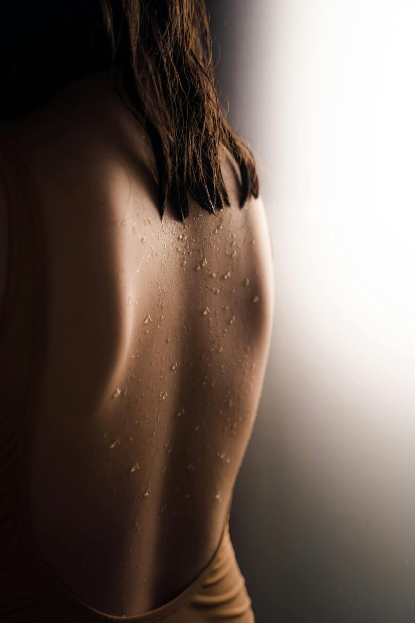 How to Keep Your Vagina Clean and Smelling Good? Close-up image of a wet back of a woman with water droplets, wearing a sleeveless top, lit by a gradient background.