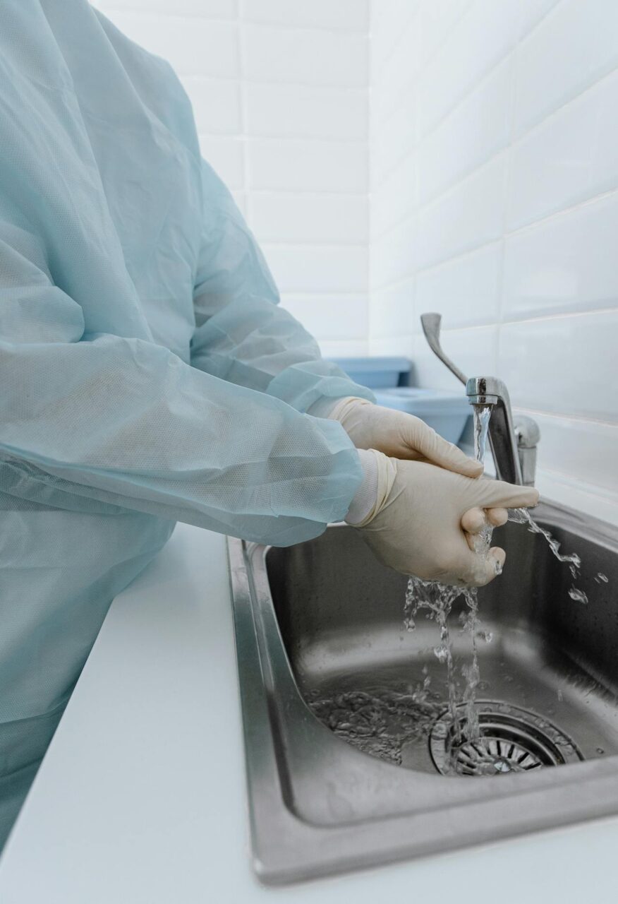 Image of a person in medical scrubs and gloves washing hands under running water at a stainless steel sink, showing uses of an antibacterial soap.