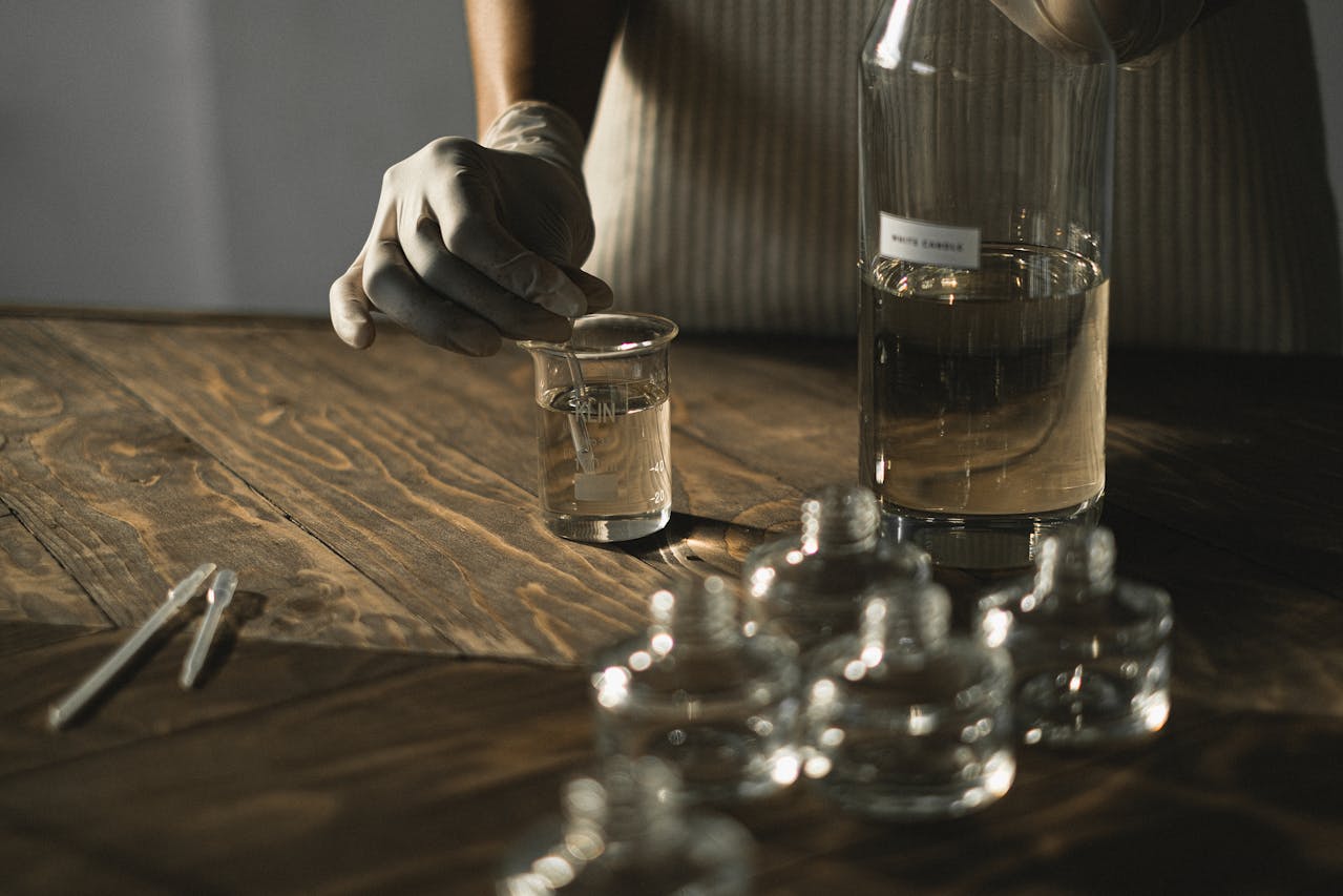 Image of a person wearing gloves mixing liquid in a beaker on a wooden table with glass bottles around, showing what is antibacterial soap good for.