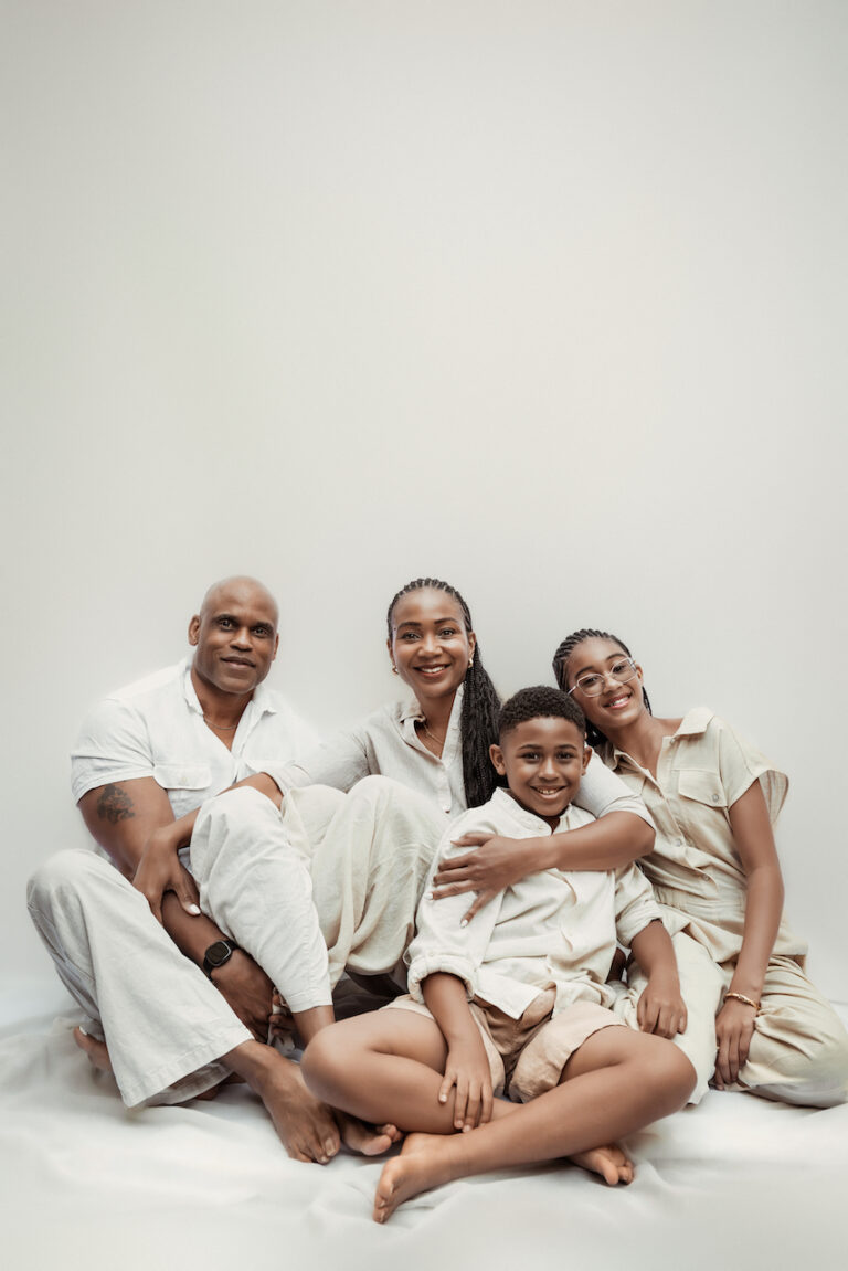 Image of a happy family of four sitting closely together on the floor, smiling against a light background, showing responsibilities of a parent.