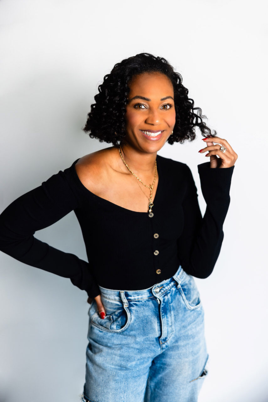 How to Break Generational Trauma: 5 Steps You Can Take Today Image of a smiling woman with curly hair wearing a black top and blue jeans, posing confidently against a white wall.
