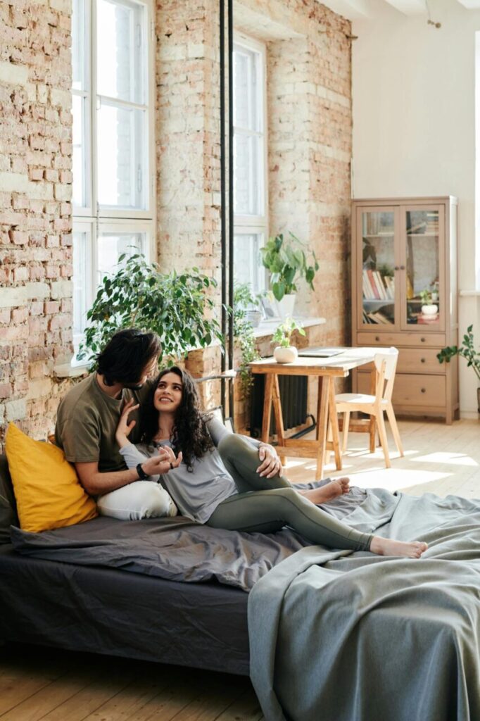 Image of a couple cuddling on a bed in a cozy loft with brick walls, plants, and natural sunlight, showing phrases to set boundaries with family.