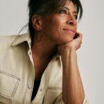 Image of a woman in beige outfit resting chin on hand, gazing thoughtfully to the side, smiling softly, showing hobbies for 60 year old woman.