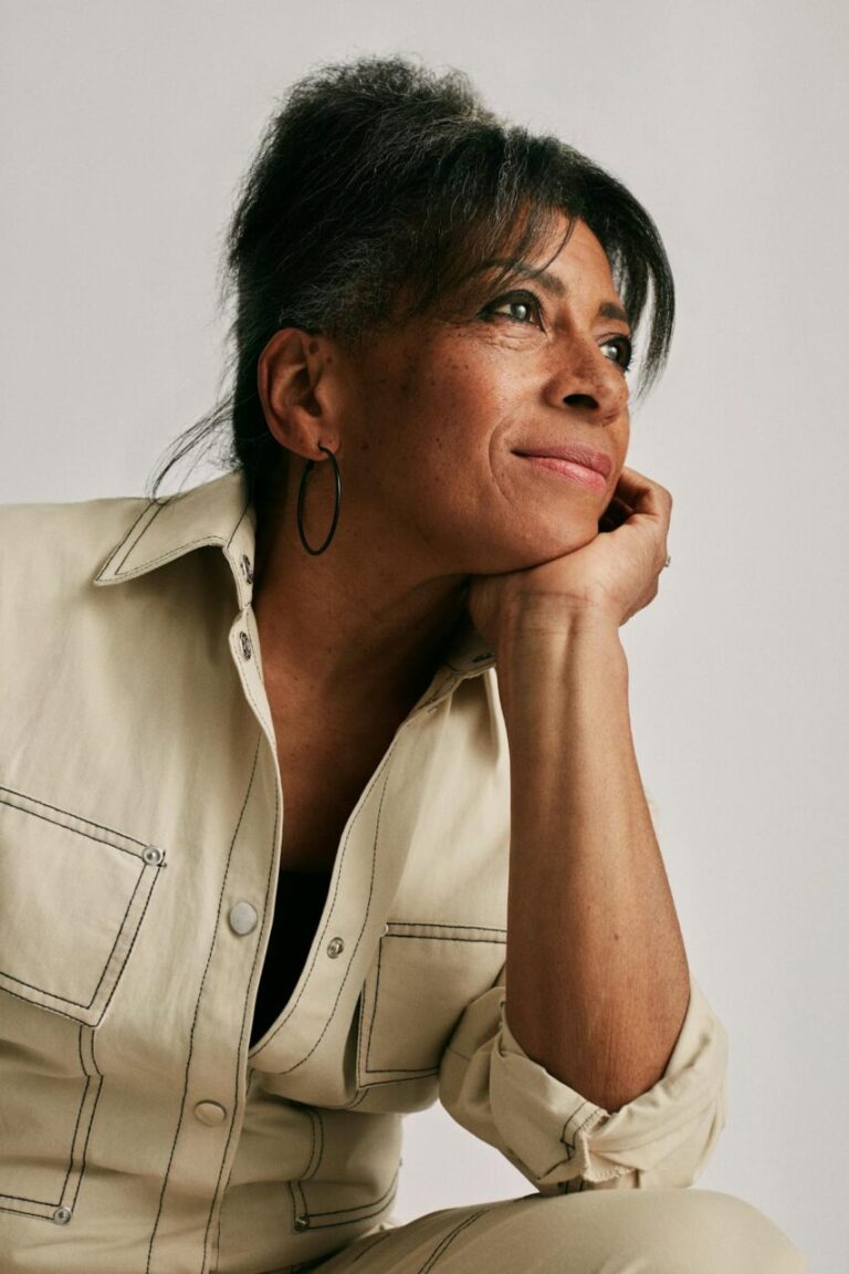 Image of a woman in beige outfit resting chin on hand, gazing thoughtfully to the side, smiling softly, showing hobbies for 60 year old woman.