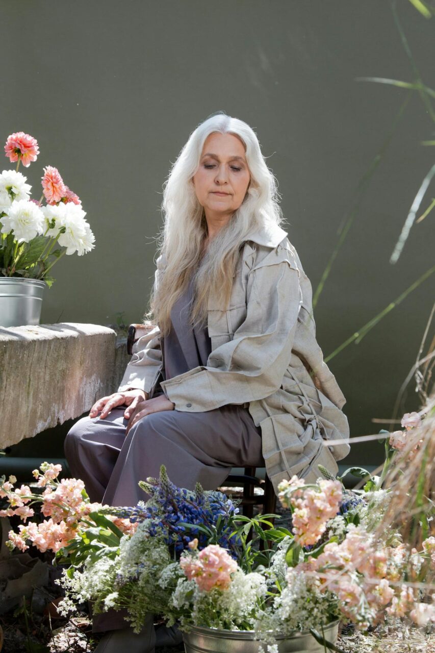 Close-up image of a elderly woman with white hair seated near colorful flower buckets in a sunlit greenhouse.