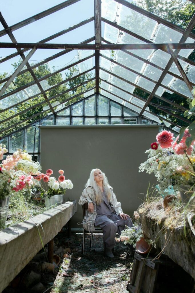 Image of a elderly woman with long white hair sits in an old greenhouse surrounded by colorful flowers, showing hobbies for 70 year old woman.
