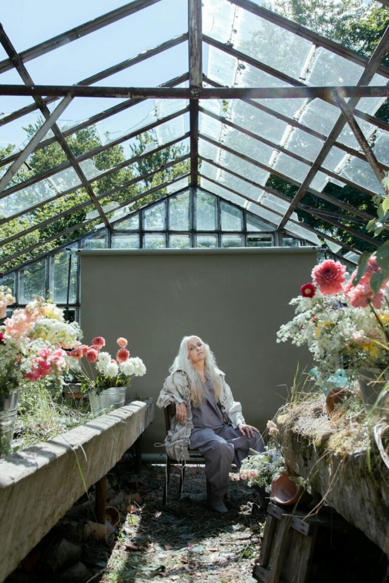 Image of a elderly woman with long white hair sits in an old greenhouse surrounded by colorful flowers, showing hobbies for 70 year old woman.