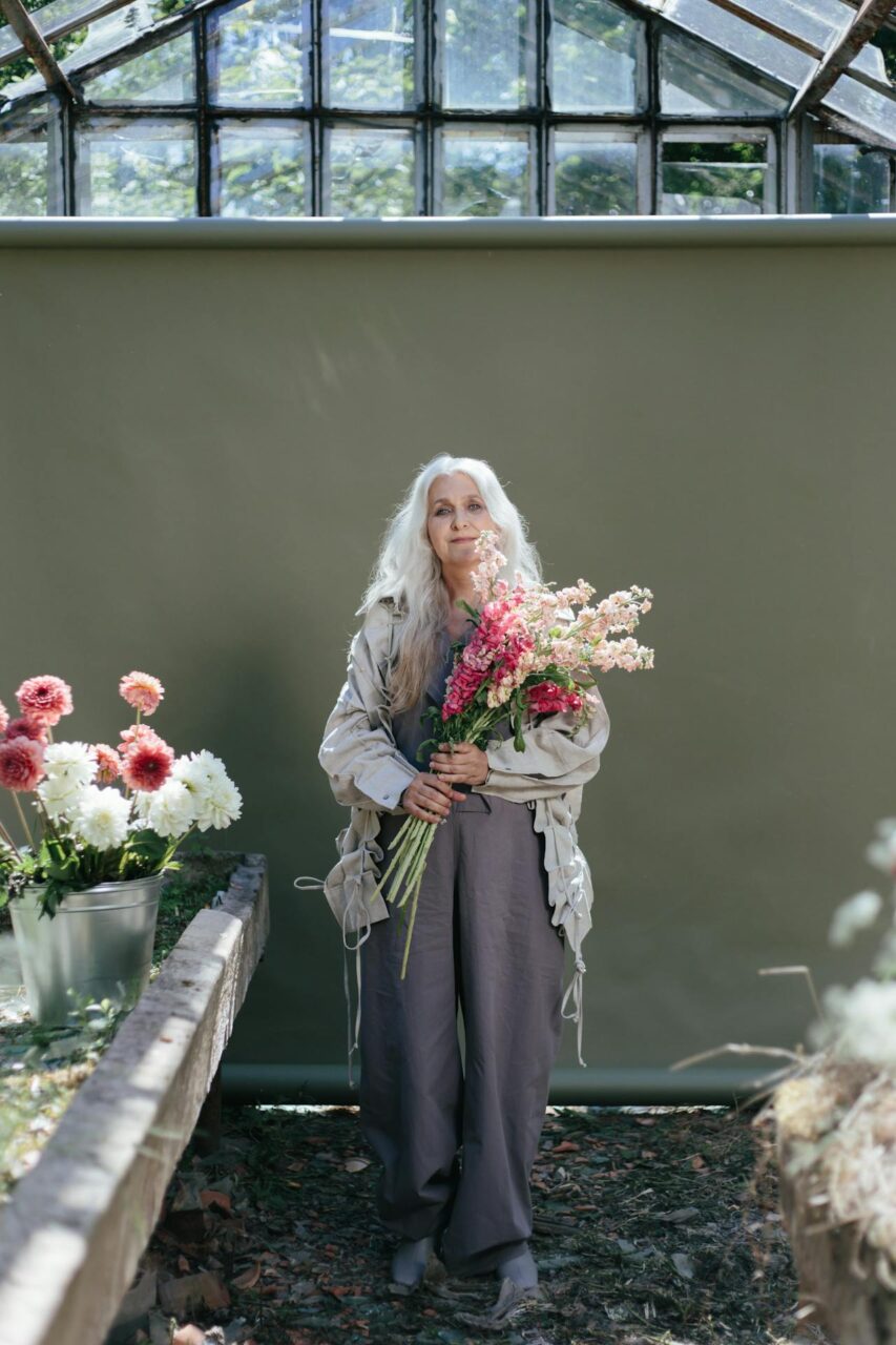 Image of a elderly woman with long white hair holding a bouquet of flowers in a greenhouse setting, showing hobbies for elderly at home.