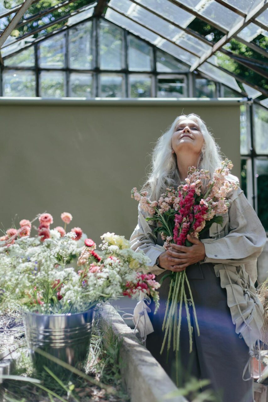 Image of a lderly woman with long white hair holds a vibrant bouquet of flowers inside a rustic greenhouse, representing hobbies for 70 year old woman.