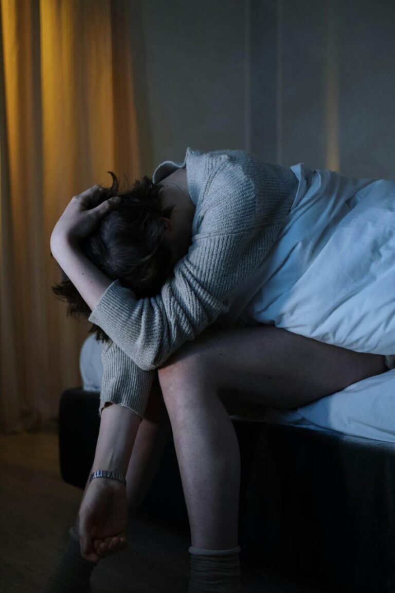 Image of a woman sitting on bed in dimly lit room, head down, arms wrapped around head, expressing distress, showing postpartum quotes.