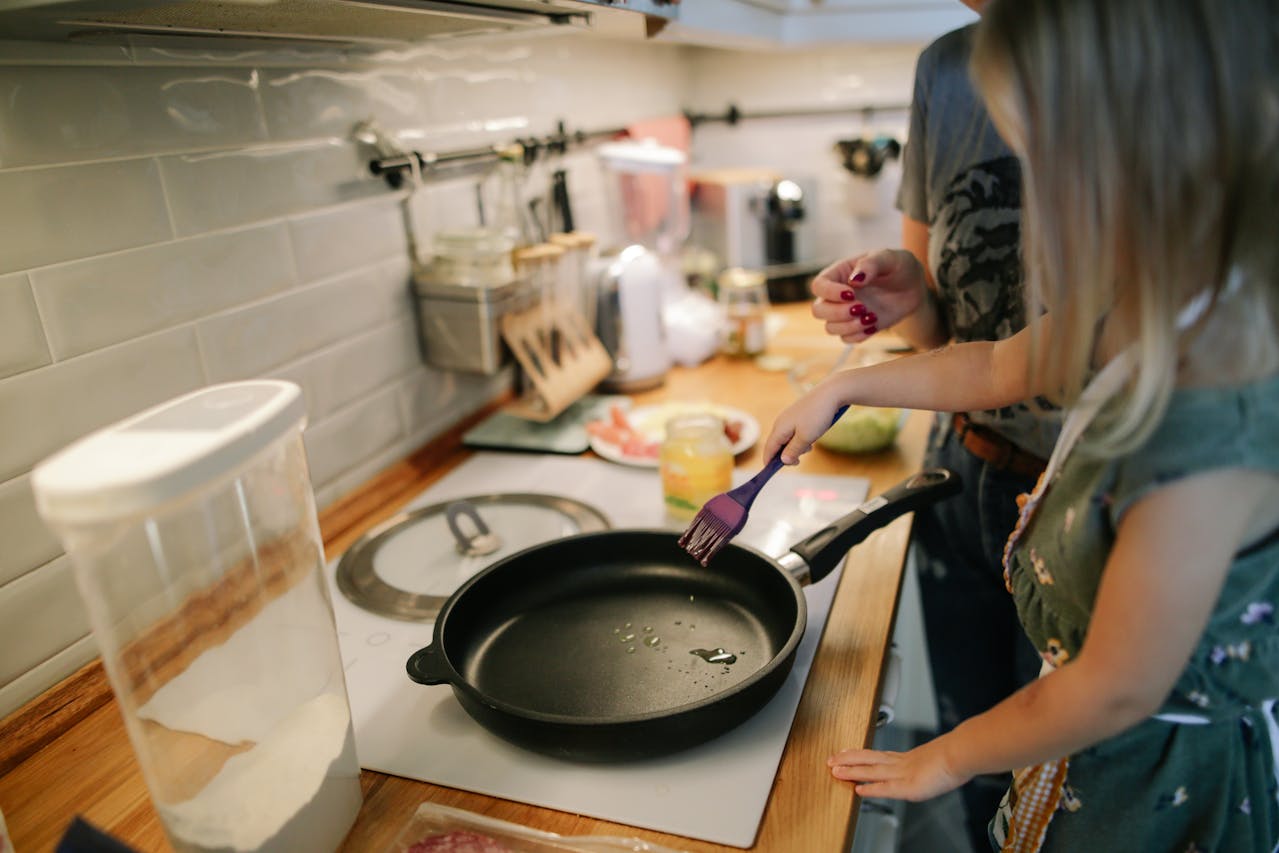 Image of a child brushing oil on a frying pan in a kitchen with an adult supervising nearby.