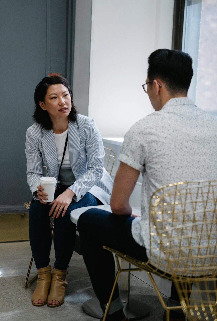 Image of a woman holding a coffee cup is engaged in a serious conversation with a man in a casual office setting.