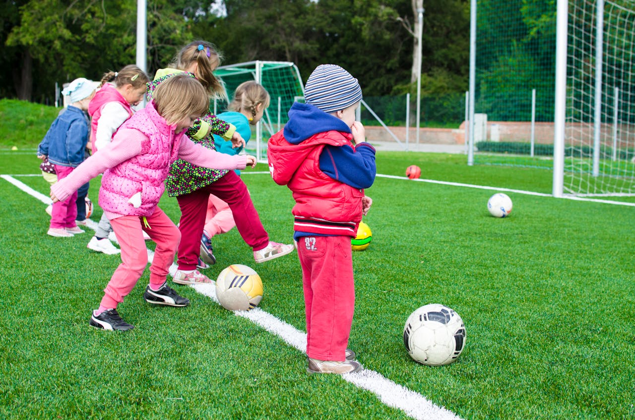 Image of a children playing soccer on a grassy field, lined up with footballs near a goalpost.