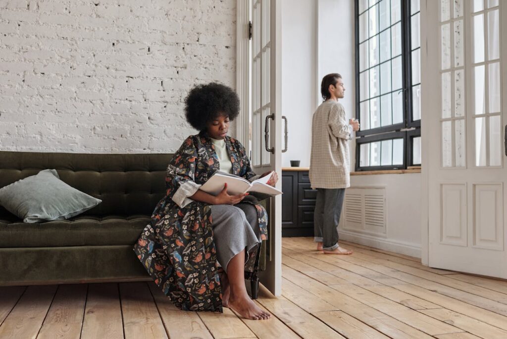Image of a woman reading book on sofa while man stands by window with coffee in cozy modern apartment, showing how to explain mom burnout to husband.