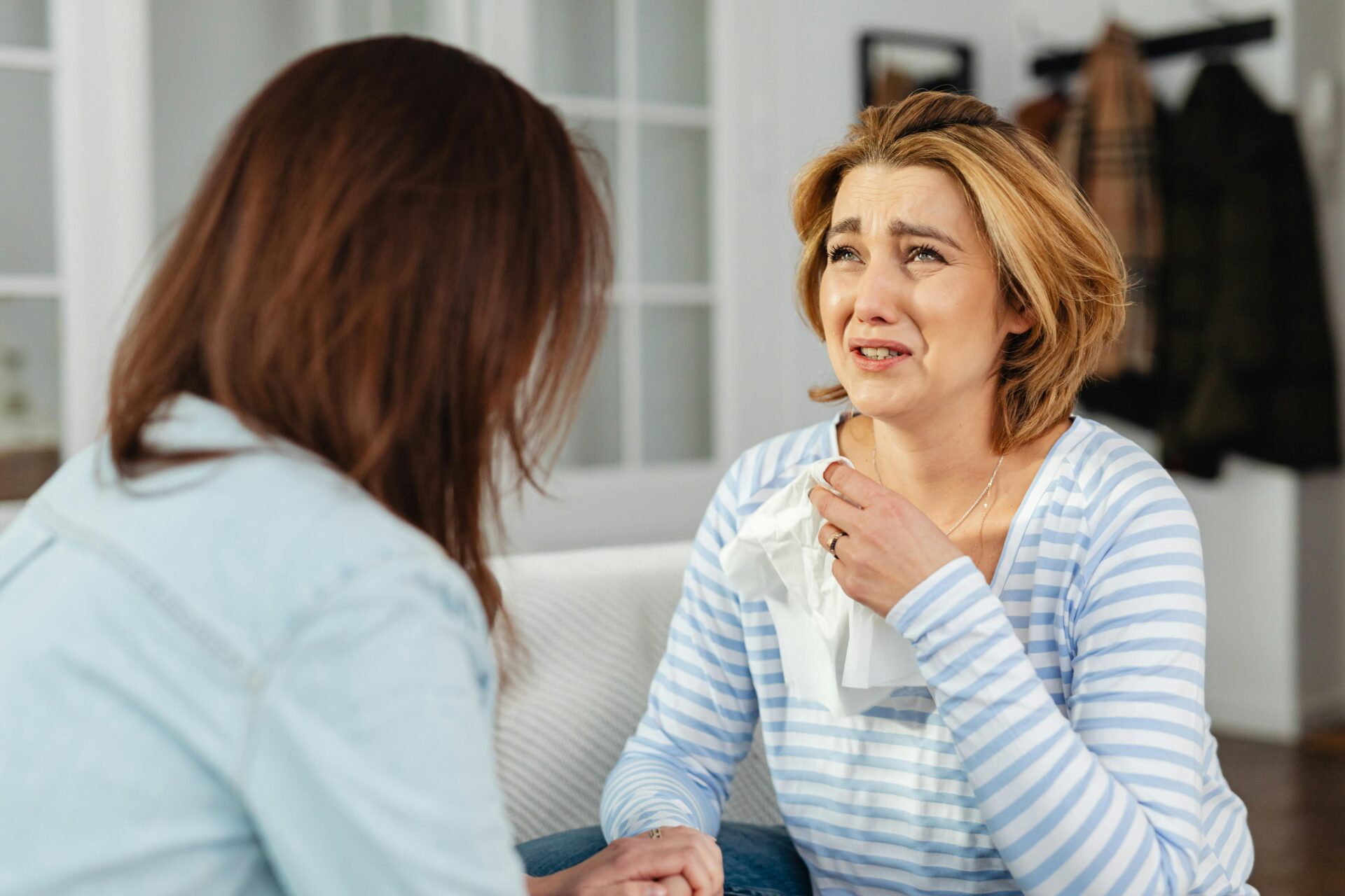 Image of a crying woman in a striped shirt holds tissues while talking to another woman who comforts her, representing how long does postpartum brain fog last.