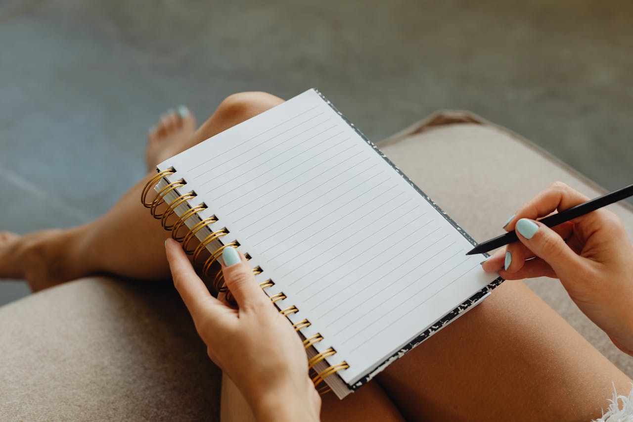 Image of a woman with painted nails holding a spiral notebook and black pencil, ready to write on blank pages.