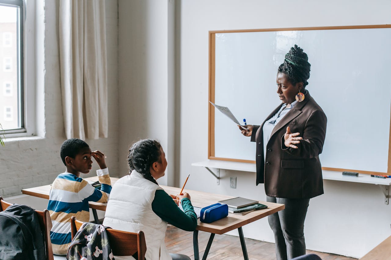 Image of a teacher holding paper while talking to two attentive students sitting at desks in a classroom.
