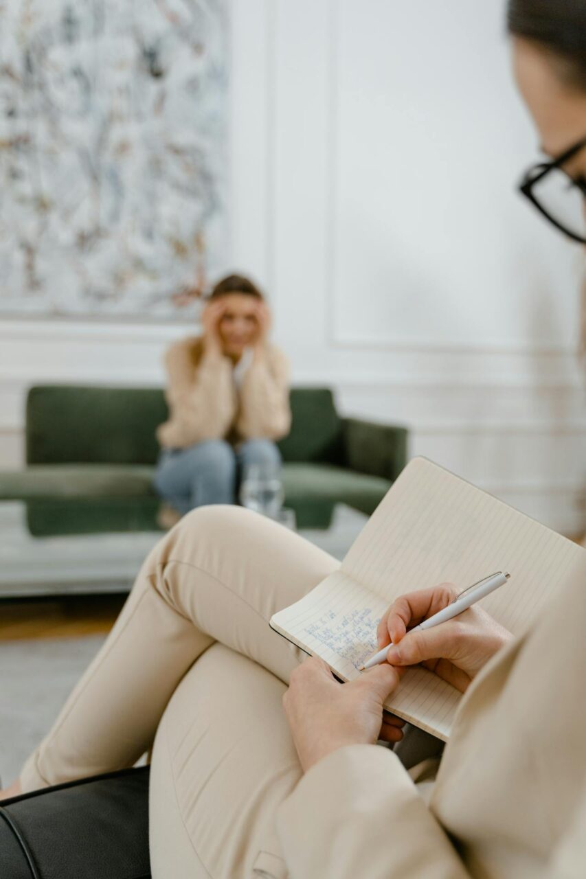 How to Break Generational Trauma: 5 Steps You Can Take Today Image of a therapist writing notes during session while a stressed woman sits on a couch in the background.