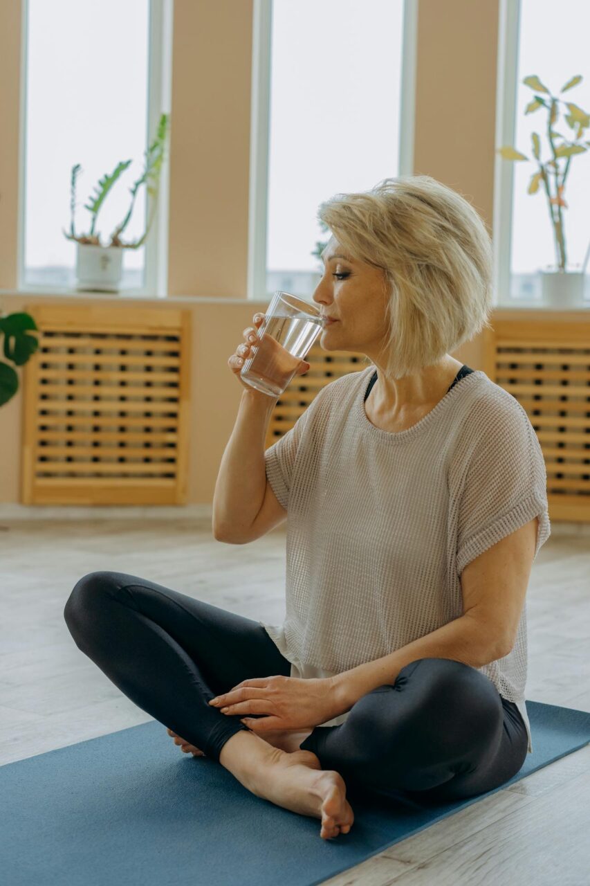 210 Best Hobbies for 60 Year Old Woman Image of a woman sitting cross-legged on yoga mat, drinking a glass of water in a bright, cozy room, showing hobbies for 60 year old woman.
