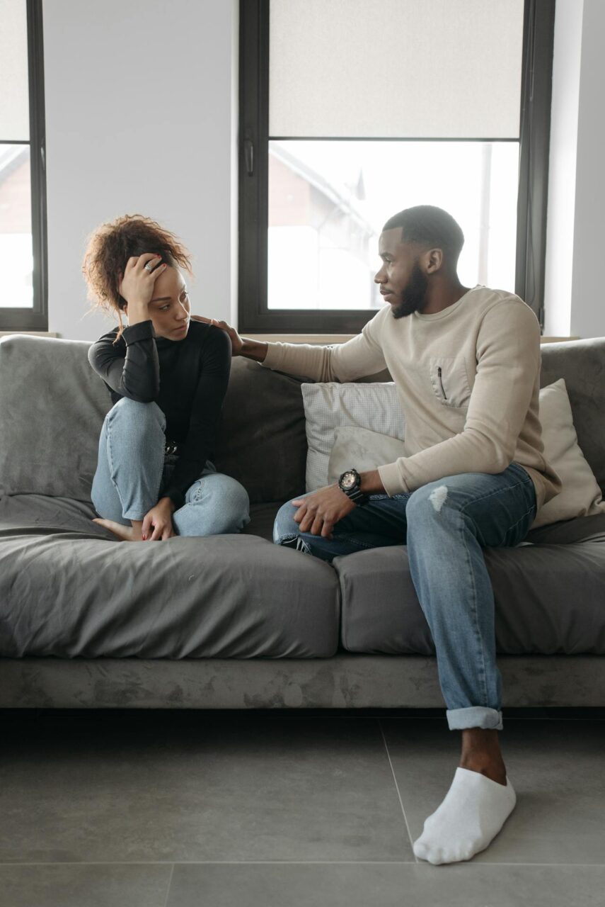 Image of a man comforting a sad woman on a gray couch in a bright living room with large windows, representing phrases to set boundaries With family.