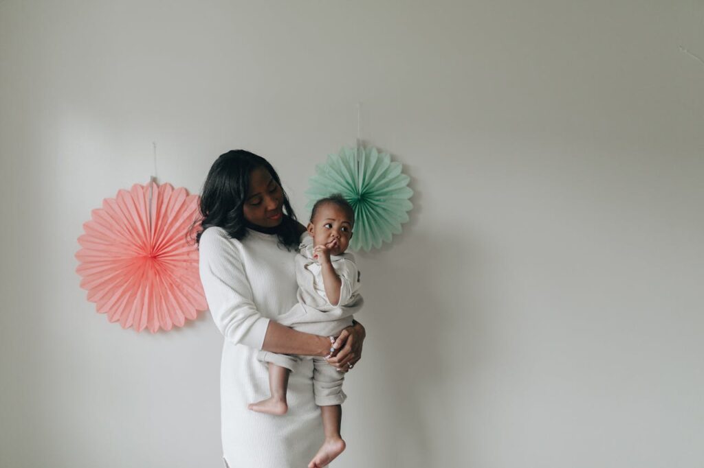 Image of a mother in white dress lovingly holds her barefoot toddler in front of pastel wall decorations.