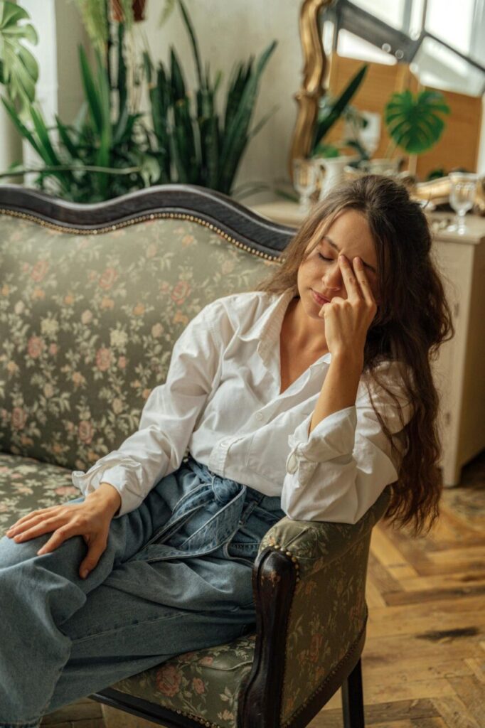 Image of a young woman in white shirt and jeans sits on floral sofa with eyes closed, resting hand on her forehead, showing how to explain mom burnout to husband.