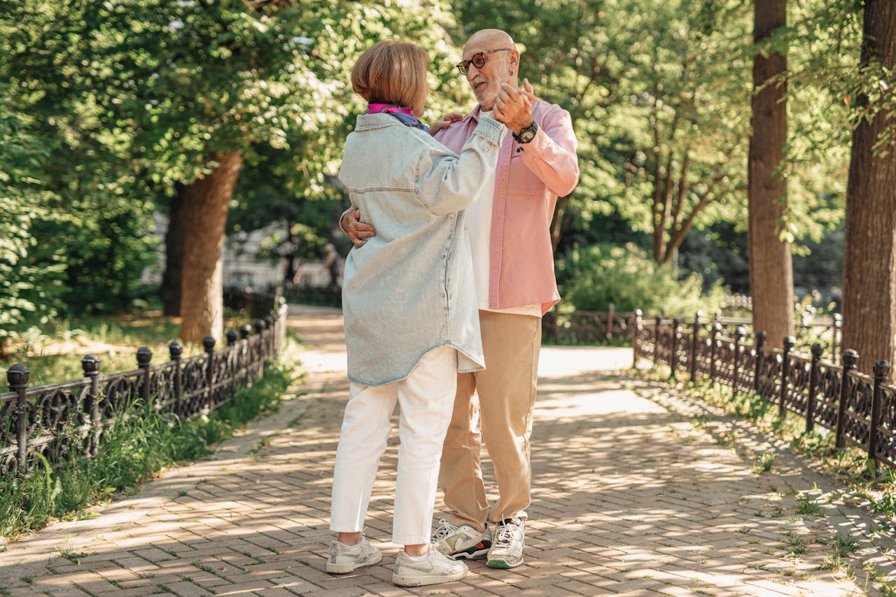 210 Best Hobbies for 60 Year Old Woman Image of a elderly couple dancing joyfully in a sunny park, surrounded by green trees and a brick walkway.