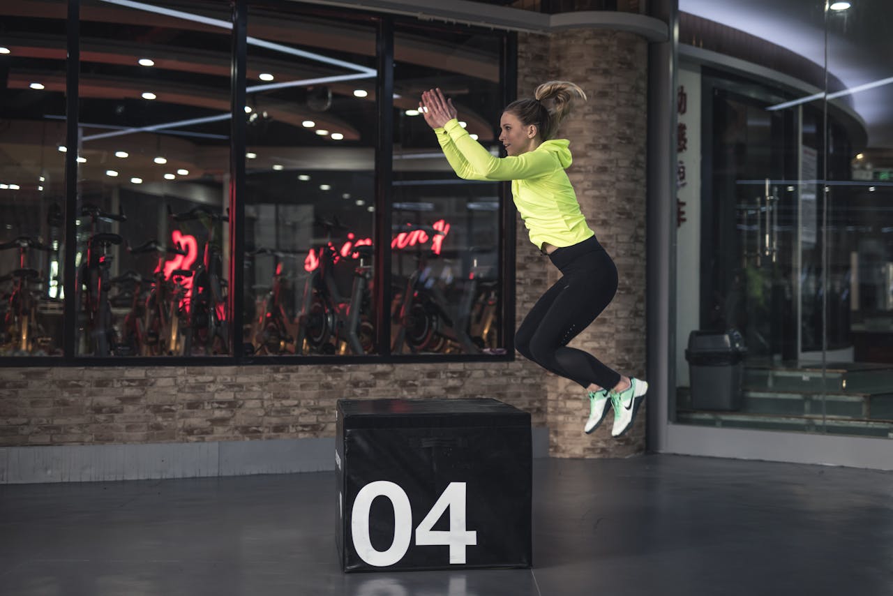 Close-up image of a woman in neon top mid-air doing box jump exercise in a gym with reflective glass walls.