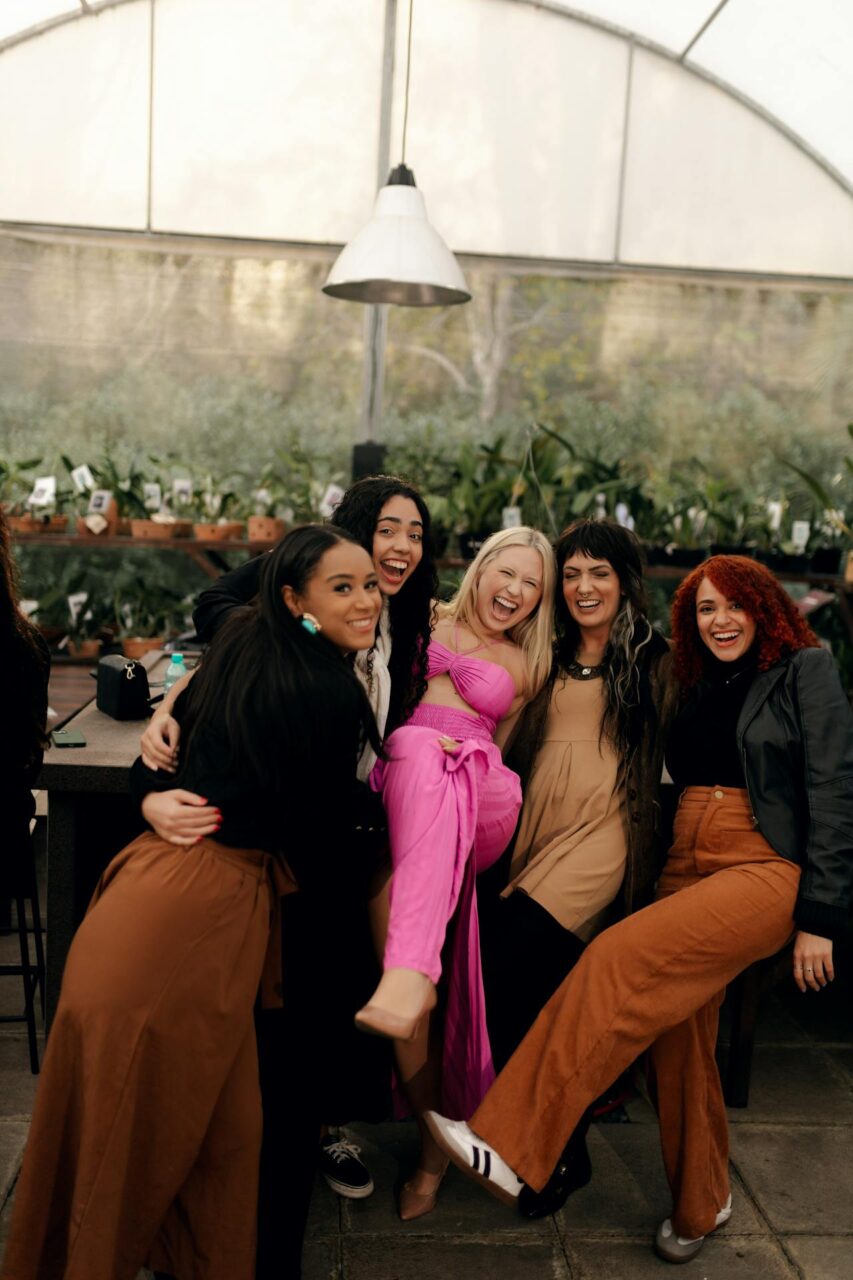 Image of a group of five joyful women posing indoors, lifting legs and smiling in a greenhouse cafe.