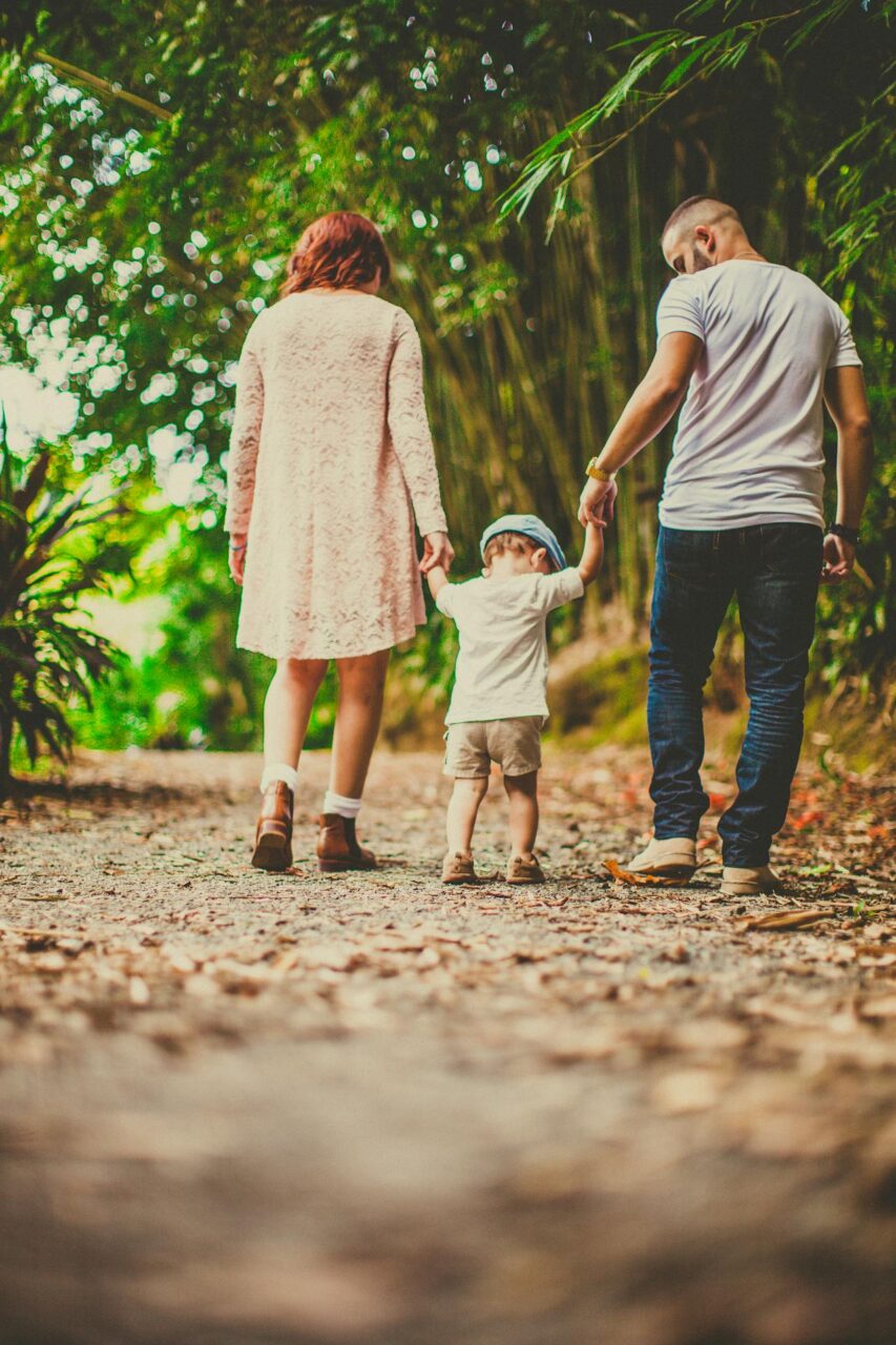 Image of a couple walks hand in hand with their toddler on a forest path surrounded by green trees and leaves.