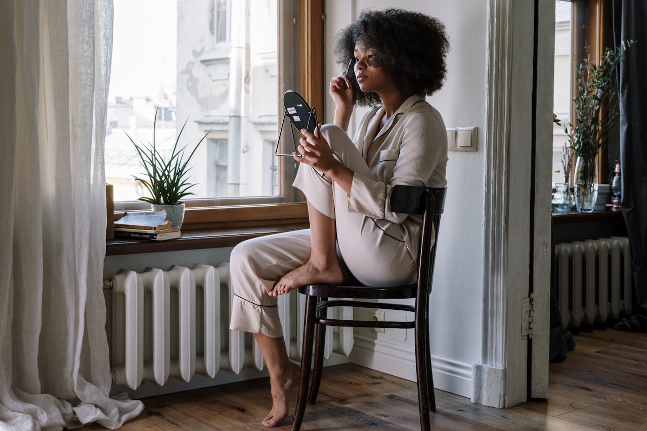 Image of a woman in pajamas sitting on a chair by a window, applying makeup with a mirror in natural light.
