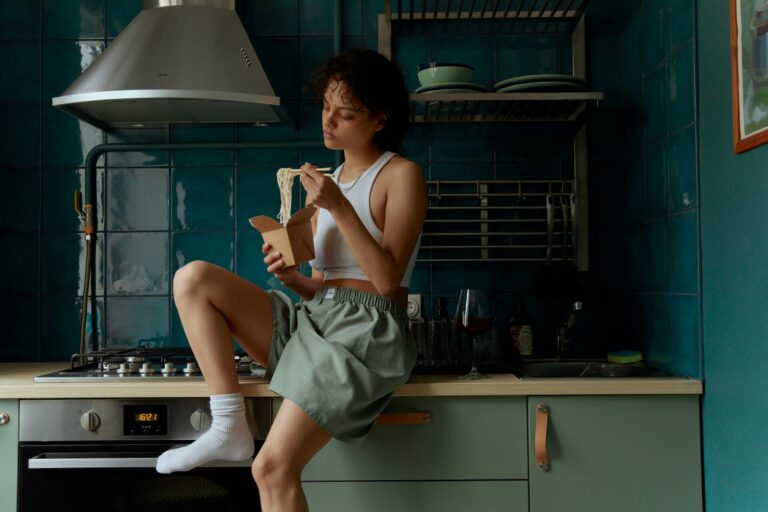 Image of a woman sitting on a kitchen counter eating noodles from a box with a fork, showing types of women's shorts.