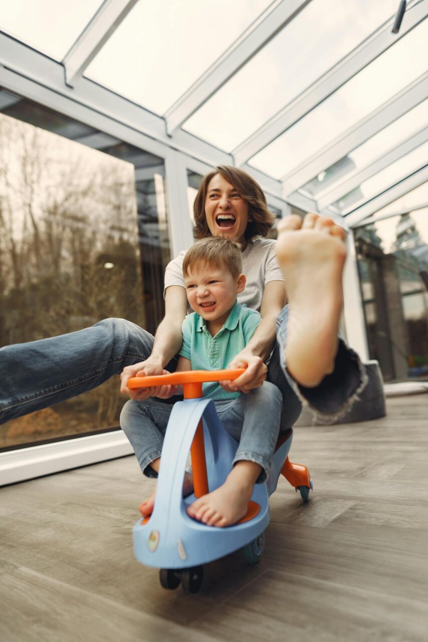 Close-up image of a joyful mother and son laugh while riding a toy scooter indoors with feet lifted in excitement, showing how to be a successful single mom.