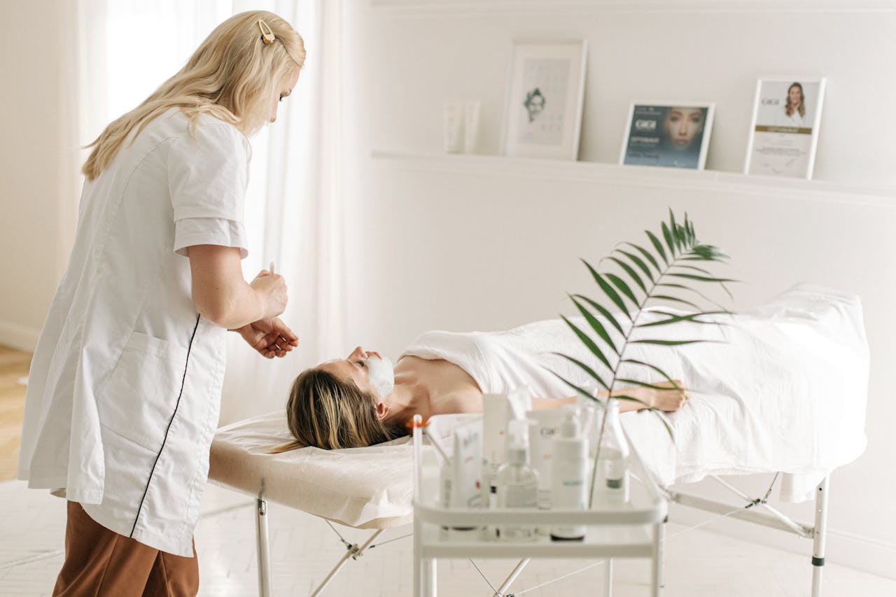 Image of a beautician applying facial treatment to a woman lying on a spa bed in a softly lit room, showing self care for single moms.