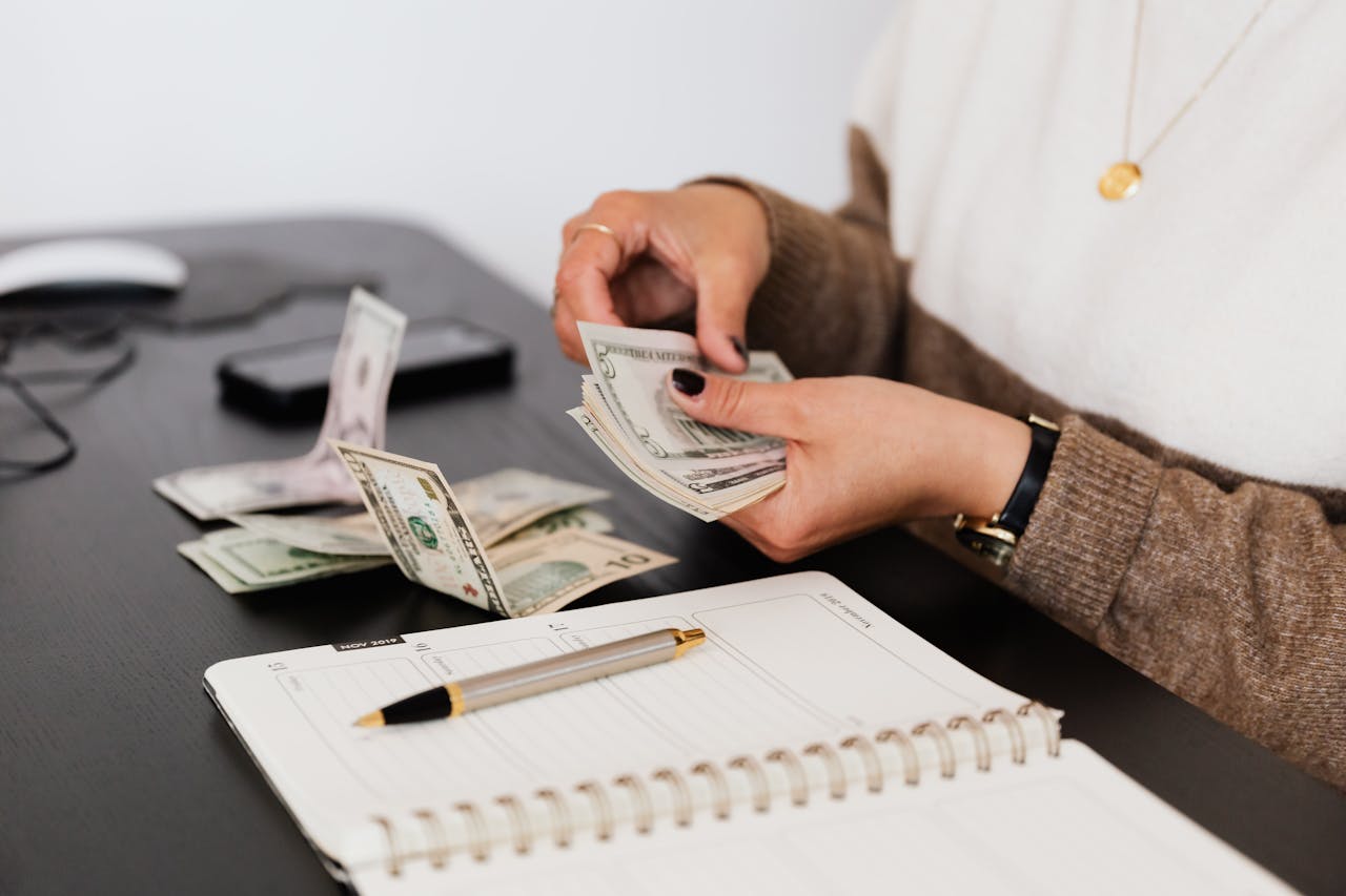 Image of a woman counts dollar bills at a desk with a notebook, pen, and smartphone beside scattered cash.