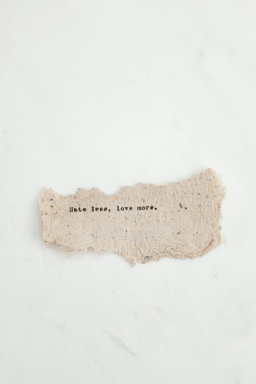 Image of a  torn piece of paper on white background with text that reads, Hate less, love more.