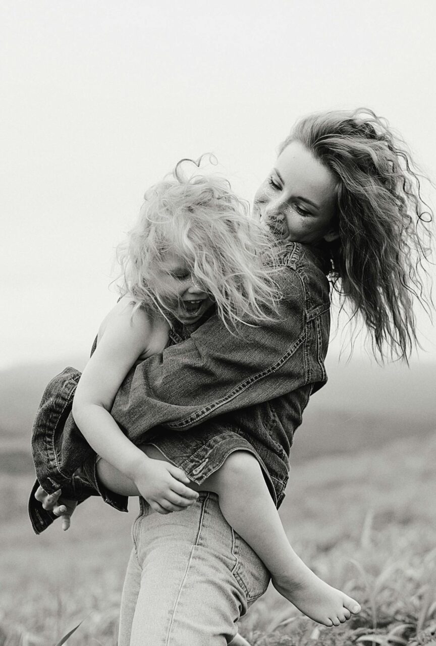 Image of a happy mother embraces her laughing daughter in a windy field, both wearing denim jackets.