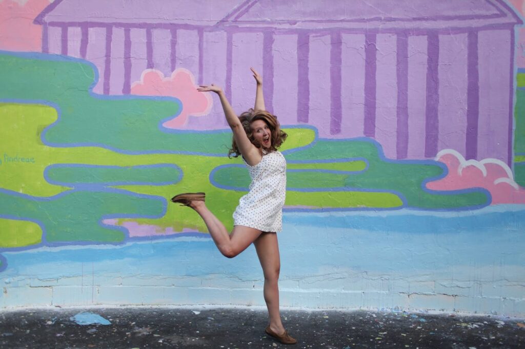 Image of a  joyful young woman in a white dress jumps playfully in front of a colorful wall mural.